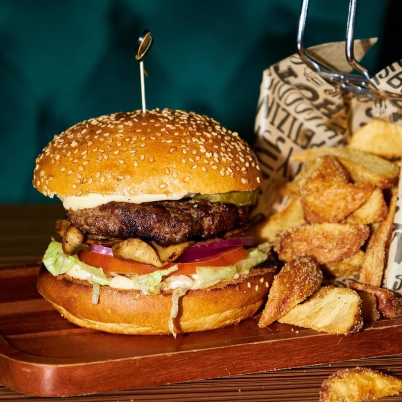 A Platan Restaurant burger with mushrooms, lettuce, tomato, onion, and a side of potato wedges.