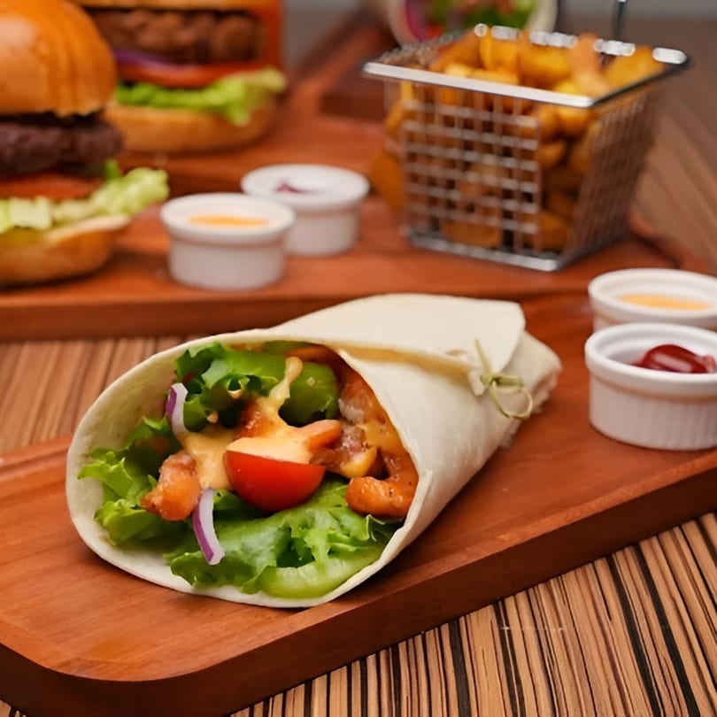 A chicken wrap with lettuce, tomato, and sauce, alongside burgers and fries on a wooden tray.