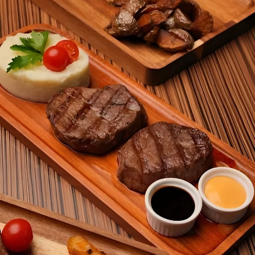 Two grilled steaks with mashed potatoes, cherry tomatoes, and two dipping sauces on a wooden board.